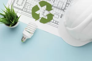 Itens relacionados à construção sustentável sobre uma planta arquitetônica: capacete de segurança branco, lâmpada fluorescente, planta verde em vaso e símbolo de reciclagem, representando práticas ecológicas na construção civil.