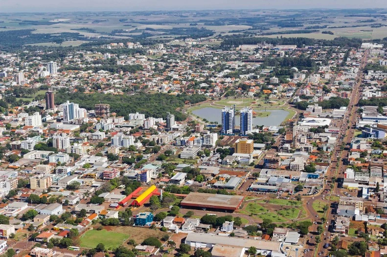 Vista aérea de Toledo Paraná, destacando áreas residenciais, comerciais e espaços públicos, com lago central cercado por prédios modernos e ruas em padrão de grade.