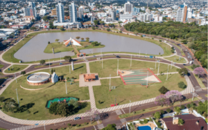 Vista aérea do Lago de Toledo com áreas de lazer, pistas de caminhada, quadras esportivas e paisagem urbana ao redor da região central da cidade.